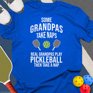Blue t-shirt with pickleball-themed text and graphics on a gray surface with pickleball equipment.